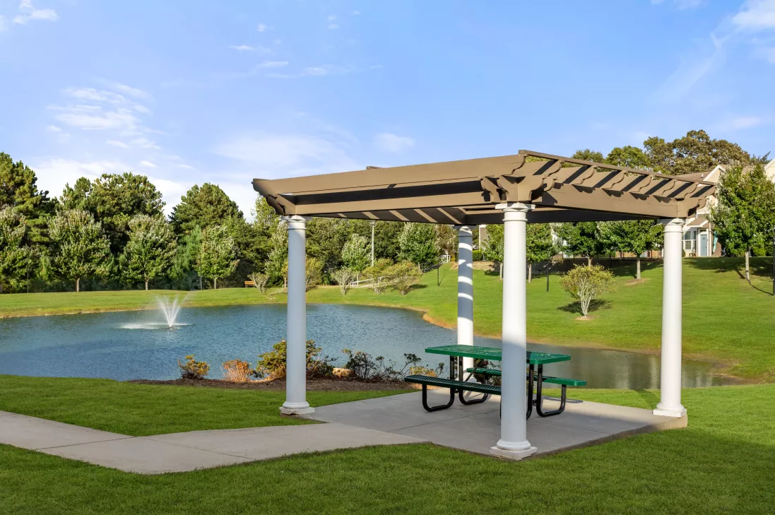 Brown pergola over a green picnic table by a pond with a fountain, surrounded by green grass and trees.