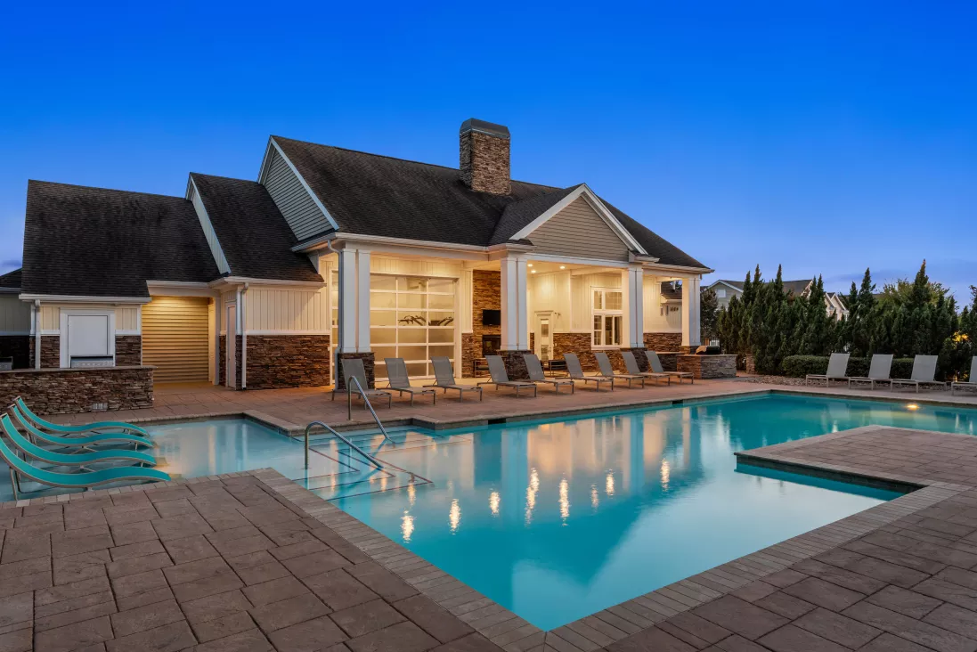 Photograph: Evening view of a brightly lit pool area with a modern clubhouse and numerous lounge chairs.