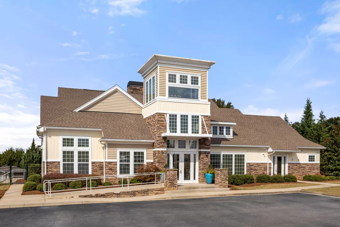 Modern clubhouse building with light yellow siding, stone accents, and a central tower under a blue sky.