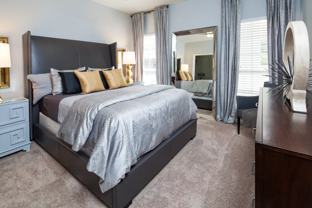 Spacious bedroom with a large bed, grey comforter, gold and black pillows, nightstand, dresser, and full-length mirror.