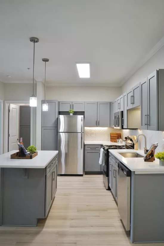 A bright, modern kitchen features gray cabinets, stainless steel appliances, white countertops, light wood floors, and an island.