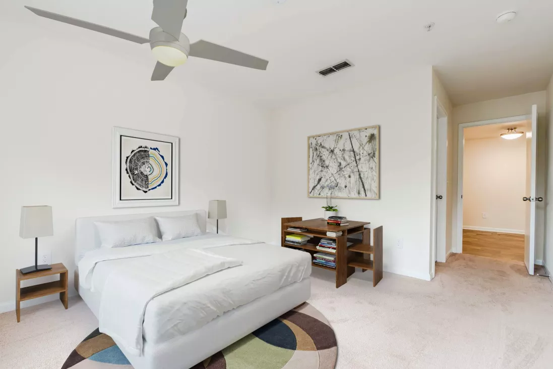 Bright bedroom with a white bed, abstract wall art, wooden desk, and a colorful round rug on light pink carpet.