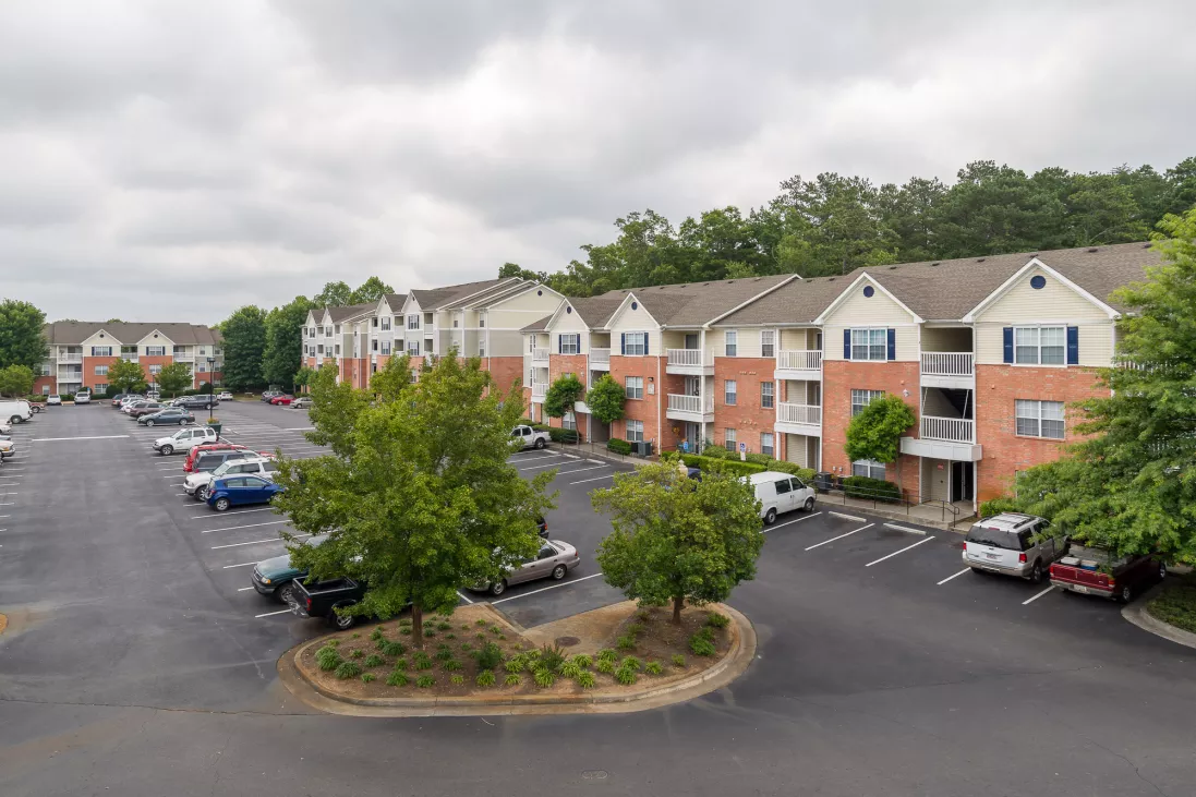 Expansive apartment complex with beige and brick buildings, a large parking lot, and green trees under a cloudy sky.